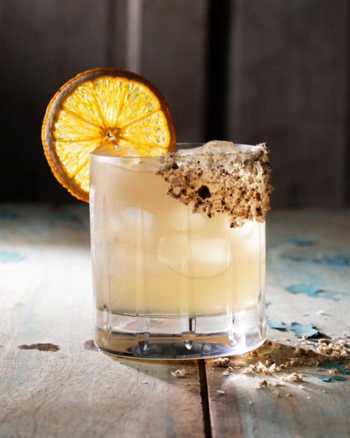 The sotoloma cocktail glass filled with a light-colored drink and ice, garnished with a salted rim and a dehydrated orange slice.