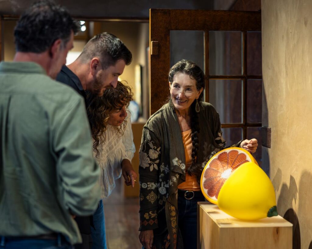 A wide shot of three people standing around a large, yellow, round sculpture of a lemon that has been cut in half to reveal a reddish-pink grapefruit interior.