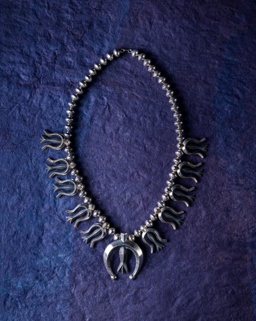 A necklace with silver beads and stylized squash blossoms, with a horseshoe-shaped naja pendant at the bottom, is laid out on a deep blue textured surface.