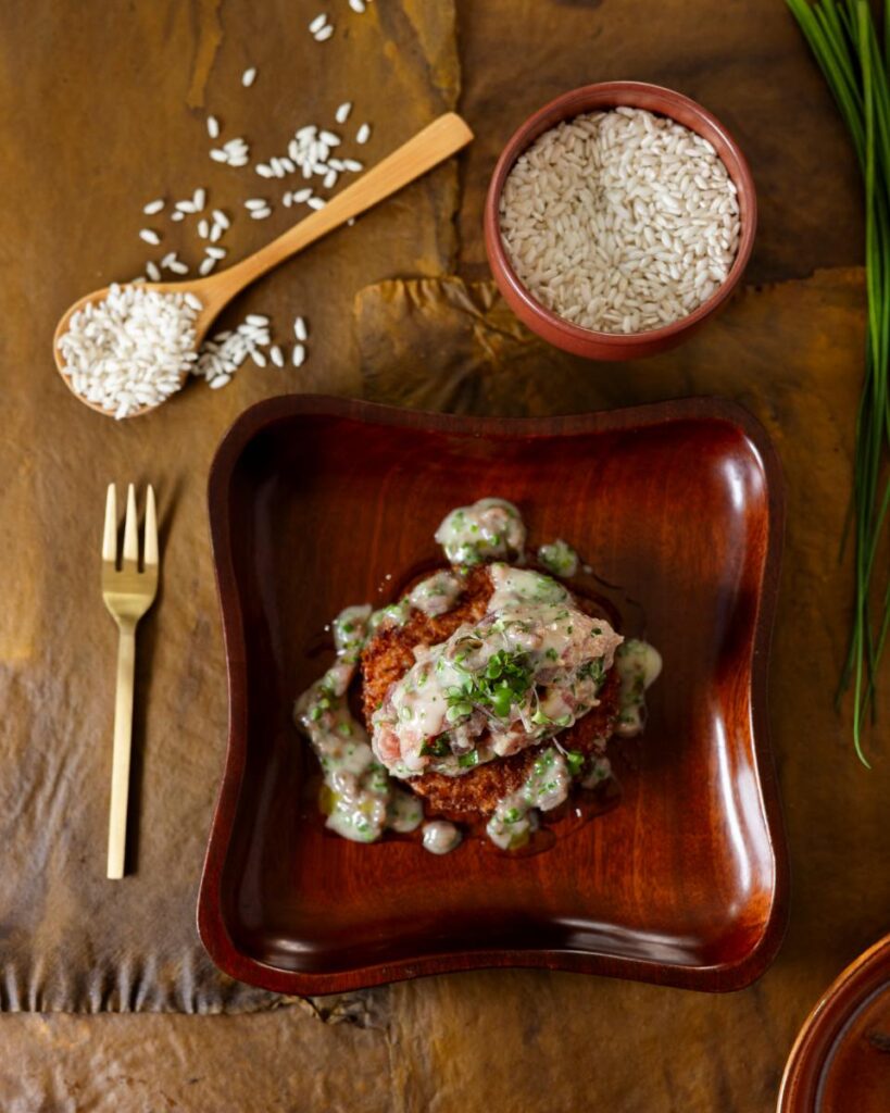A crispy risotto cake topped with tuna tartare, herbs, and a cream sauce is on a square wooden plate.