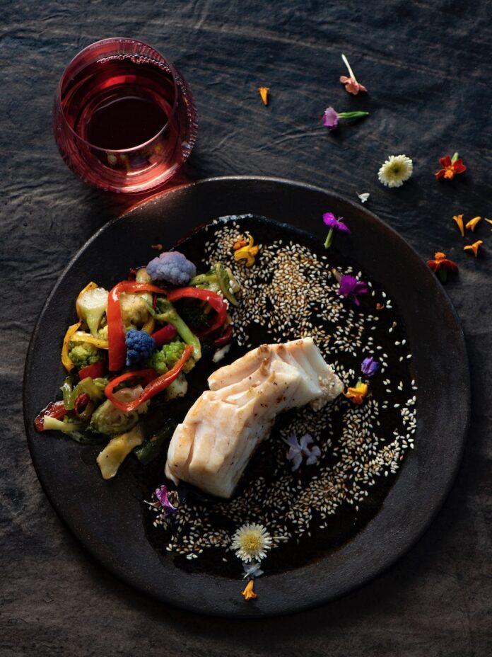 Halibut in Salsa Culiché with seeds and flowers on black plate.