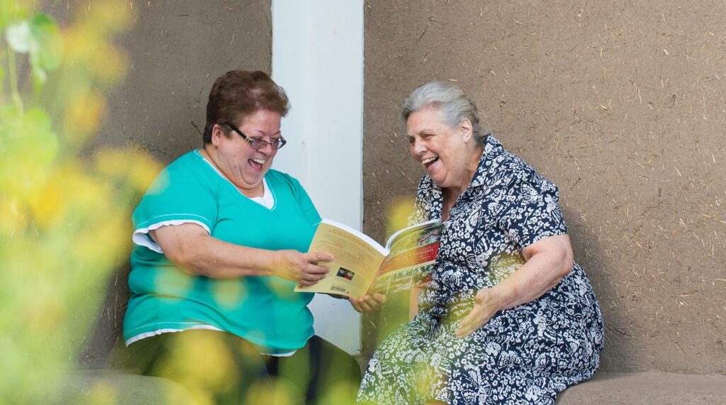 A woman in a pattern dress sits with a woman in blue, looking over a book.
