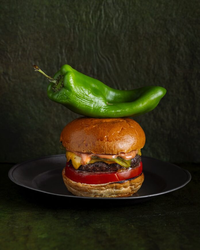 Green Chile Cheeseburger TABLE Magazine A cheeseburger sits on a plate with a large green chile sitting on top of it.