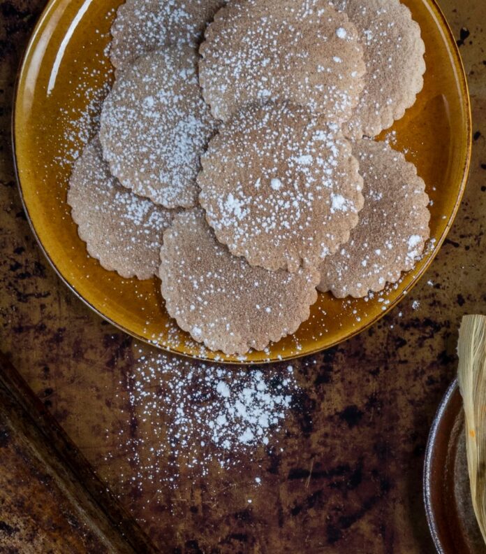 Happy Biscochitos TABLE Magazine A brown platter of round, scalloped-edged biscochito cookies dusted with powdered sugar, with some sugar sprinkled on the wooden table beside the platter.