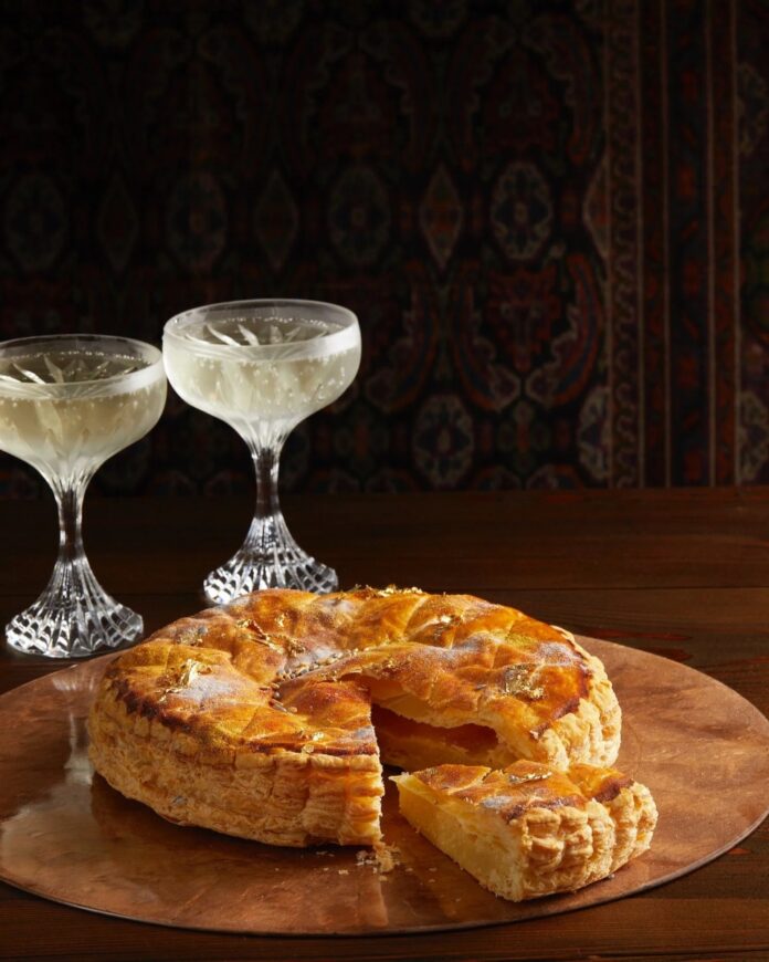 King Cake served on a round, flat, brownish plate with a piece cut and protruded out. Two stylish glasses filled with a drink.