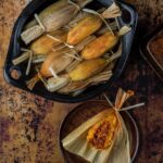 A black bowl filled with traditional tamales wrapped in corn husks, with one unwrapped tamale on a smaller plate showing a rich, red filling.
