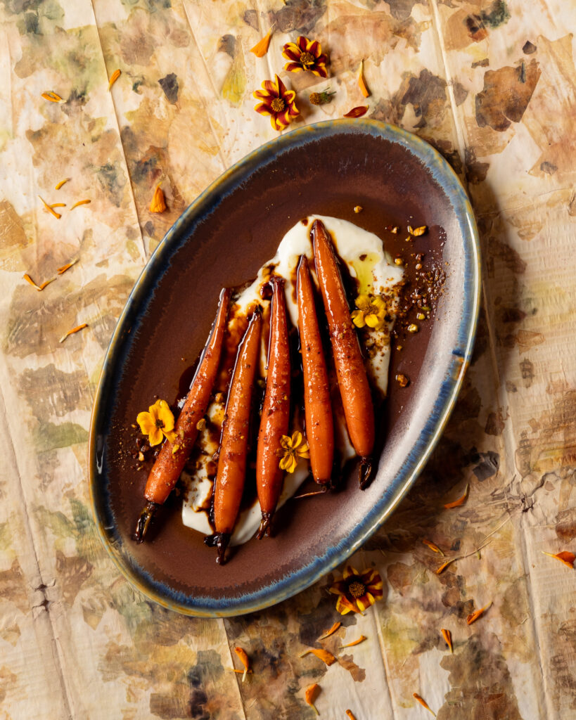 Carrots on a brown plate on top of a white sauce with yellow flowers around the. 