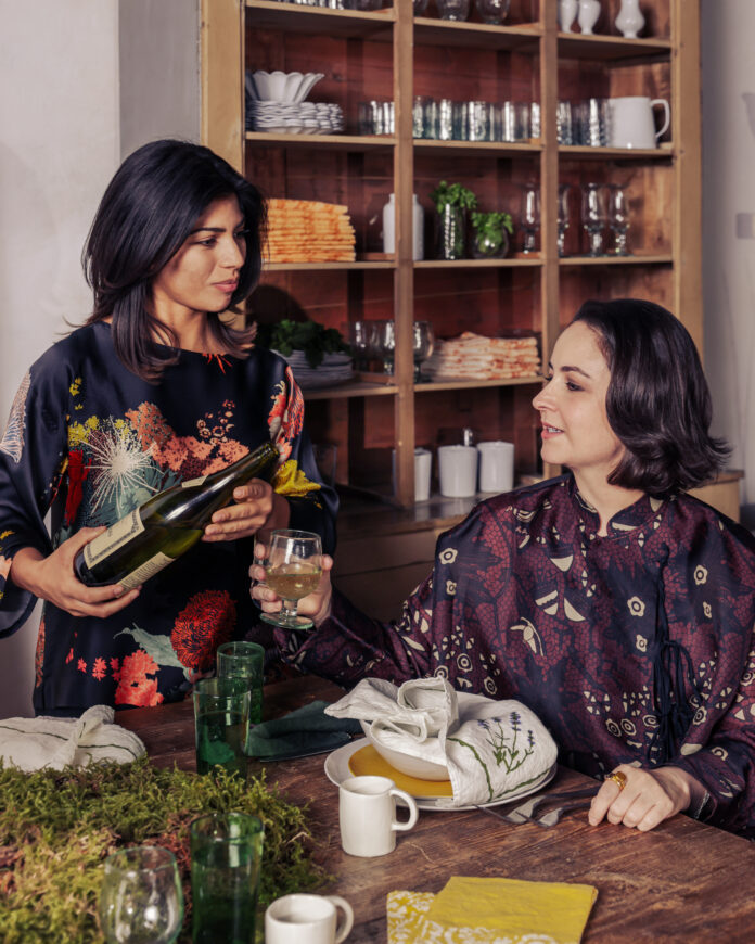 Two women sit at a wooden table with plating all around it.