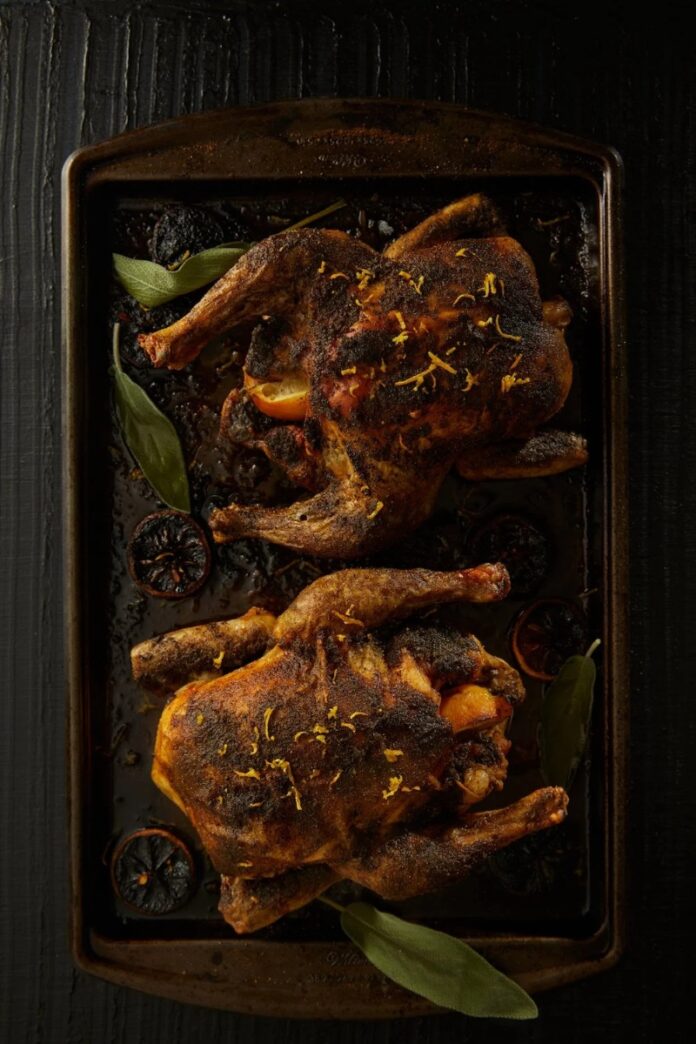 Two crispy roasted lemon chickens seasoned with sage, garnished with lemon zest and sage leaves on a dark baking sheet.