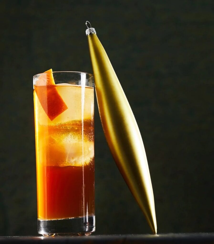 A rocks glass of drunk uncle cocktail with a large ice cube and an orange peel, next to a gold Christmas ornament.
