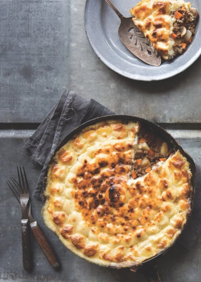 A cast iron skillet filled with Shepherd's pie topped with a golden-brown, bubbly rutabaga and potato mash, with a serving on a small gray plate.