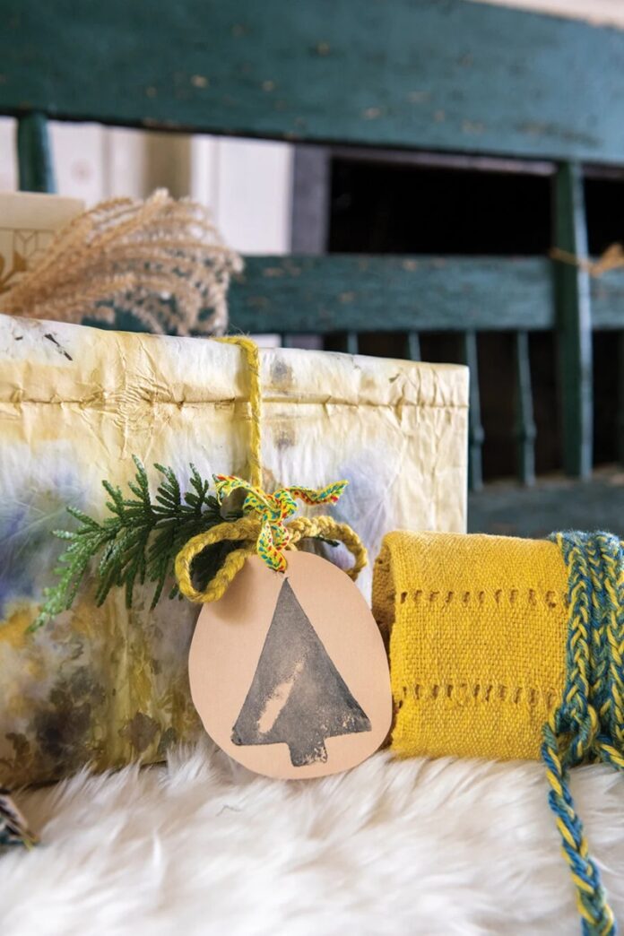 An eco-friendly wrapped gift, tied with twine and a natural paper tag with a stamped Christmas tree, next to a woven yellow fabric roll.