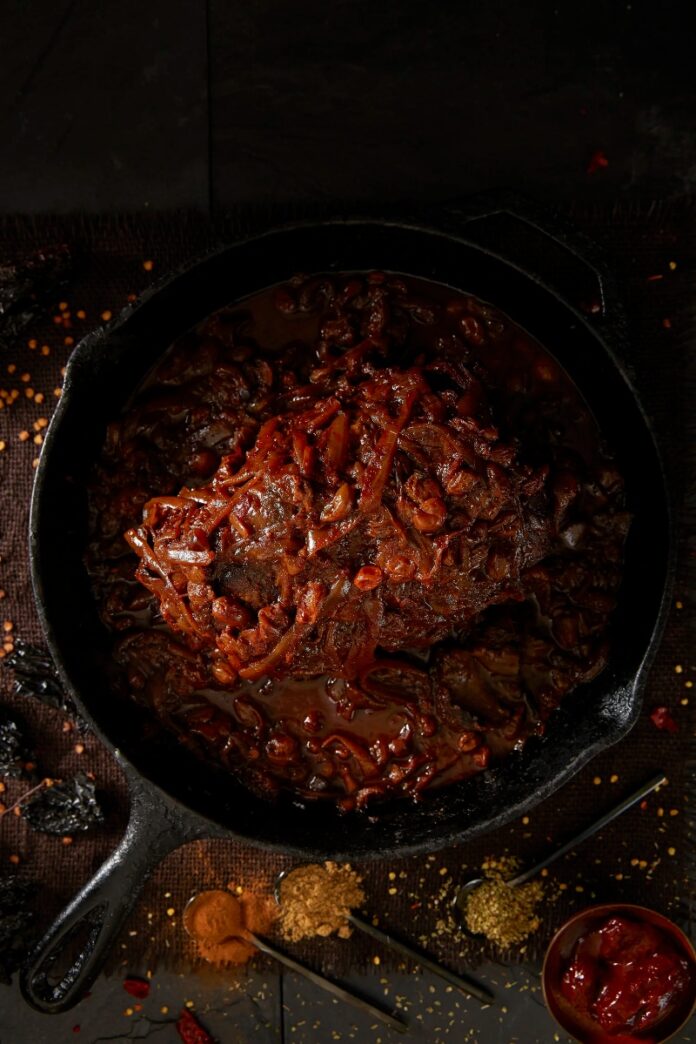 A cast iron pan filled with wild boar ragu, with various spices and peppers scattered on a dark surface around the pan.