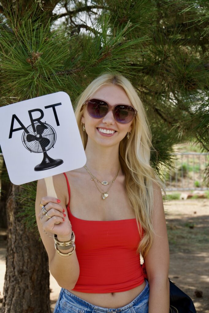 Keeping cool at IFAM, 7/11/25_Crosby A woman in a red tank top holds up an art fan sign.