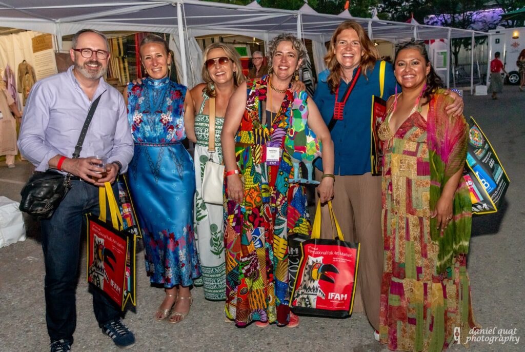 International Folk Art Market 2025 TABLE Magazine 5 A group of people stand in colorful clothes amongst artist booths with bags in hand.