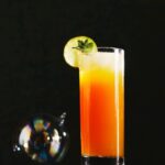 A tall glass of an orange and yellow kid's choice cocktail, garnished with a lime slice and mint, sits on a dark surface.
