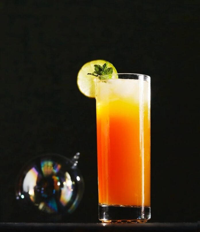 A tall glass of an orange and yellow kid's choice cocktail, garnished with a lime slice and mint, sits on a dark surface.