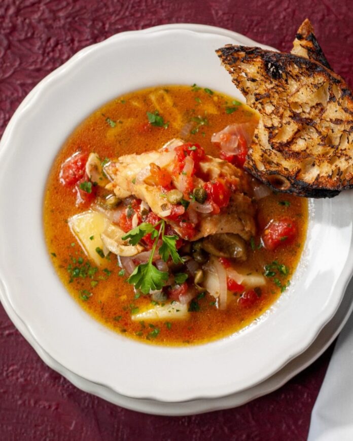 A white bowl filled with Baccalà fish stew with potatoes and tomatoes, served with a piece of grilled, charred bread on the rim.