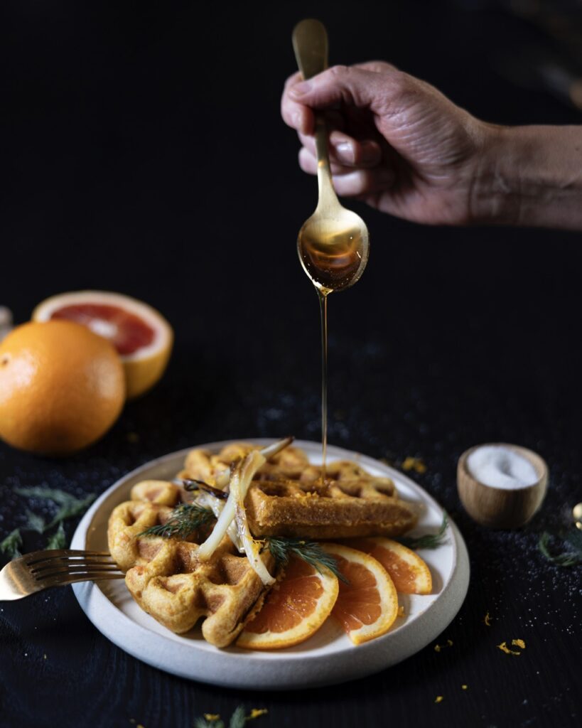 A person drizzles honey over a waffle with orange slices on the plate too.