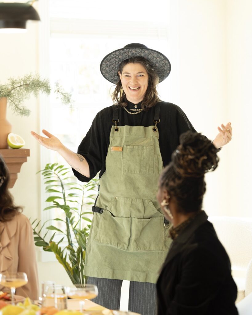 A woman with a green apron and hat talks to a dinner table.