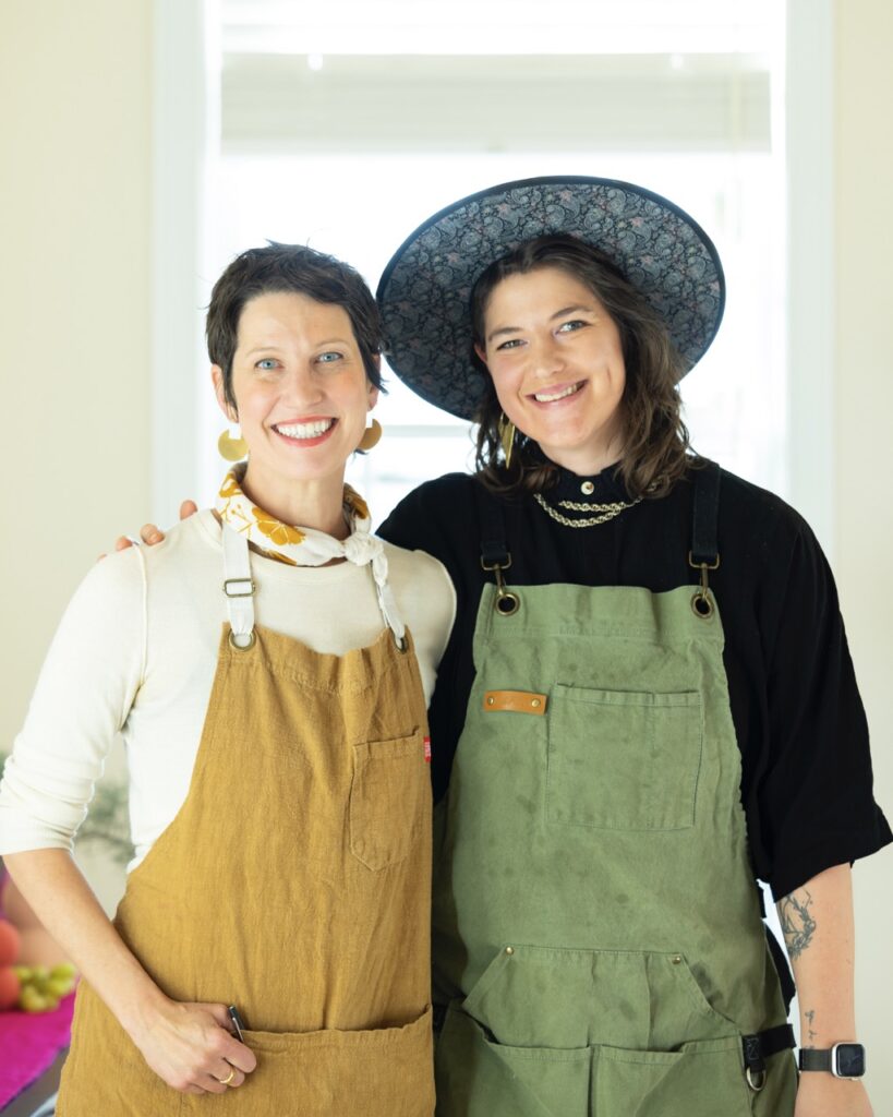 A woman in a mustard apron and a woman in a sage green apron stand together.