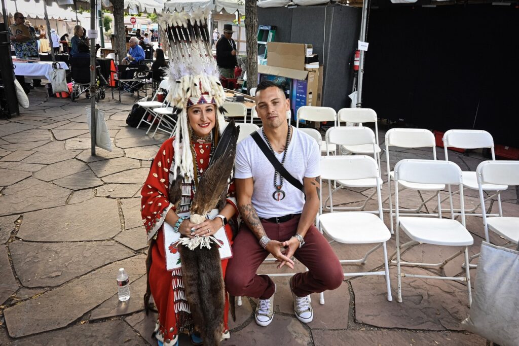 Santa Fe Indian Market 2025 Recap TABLE Magazine 3 A person in native dress and another man sit on chairs together at a festival.