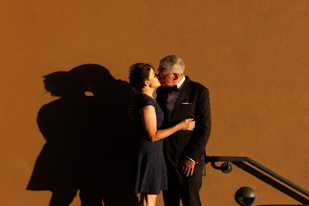 Audience Experience Portrait Series July 4 2025 Santa Fe Opera A woman in a black dress and a man in a black suit kiss in front of a brown wall.