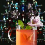 A glass mug of peacekeeper cocktail, garnished with a rose and a lemon peel, sits in front of a blurry, colorful background.