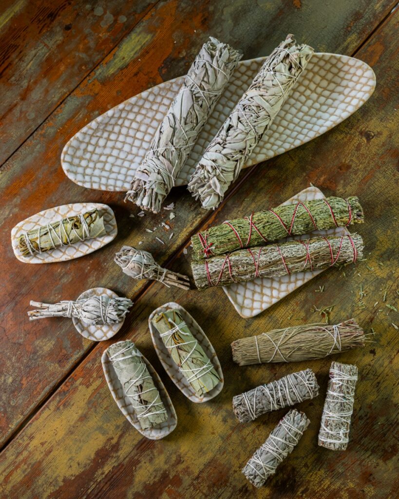A collection of white sage and cedar aromatic smudge sticks resting on handmade glazed ceramic plates on a distressed wooden surface.