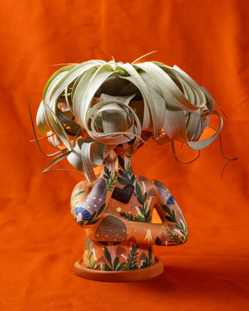 Whimsical ceramic planter shaped like a person's torso, decorated with mushrooms and flowers, holding a curly air plant on its head against an orange background.