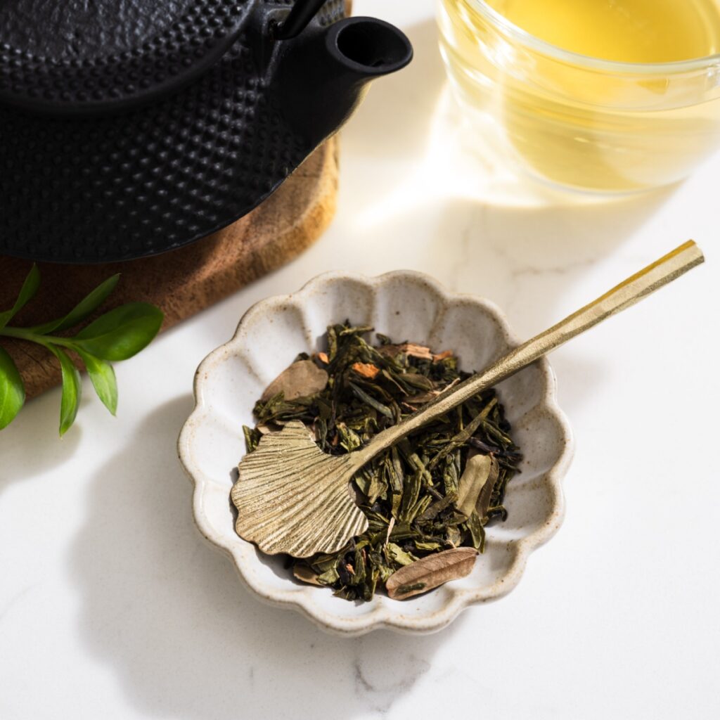 A gold-finished stainless steel ginkgo leaf spoon resting in a scalloped bowl of loose green tamarind lychee tea, next to a black cast iron teapot.
