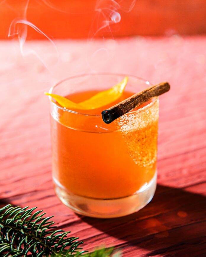 A Biscochito Old Fashioned cocktail in a rocks glass with a cinnamon sugar rim, orange peel, and smoking cinnamon stick, on a red wooden surface with pine branches.