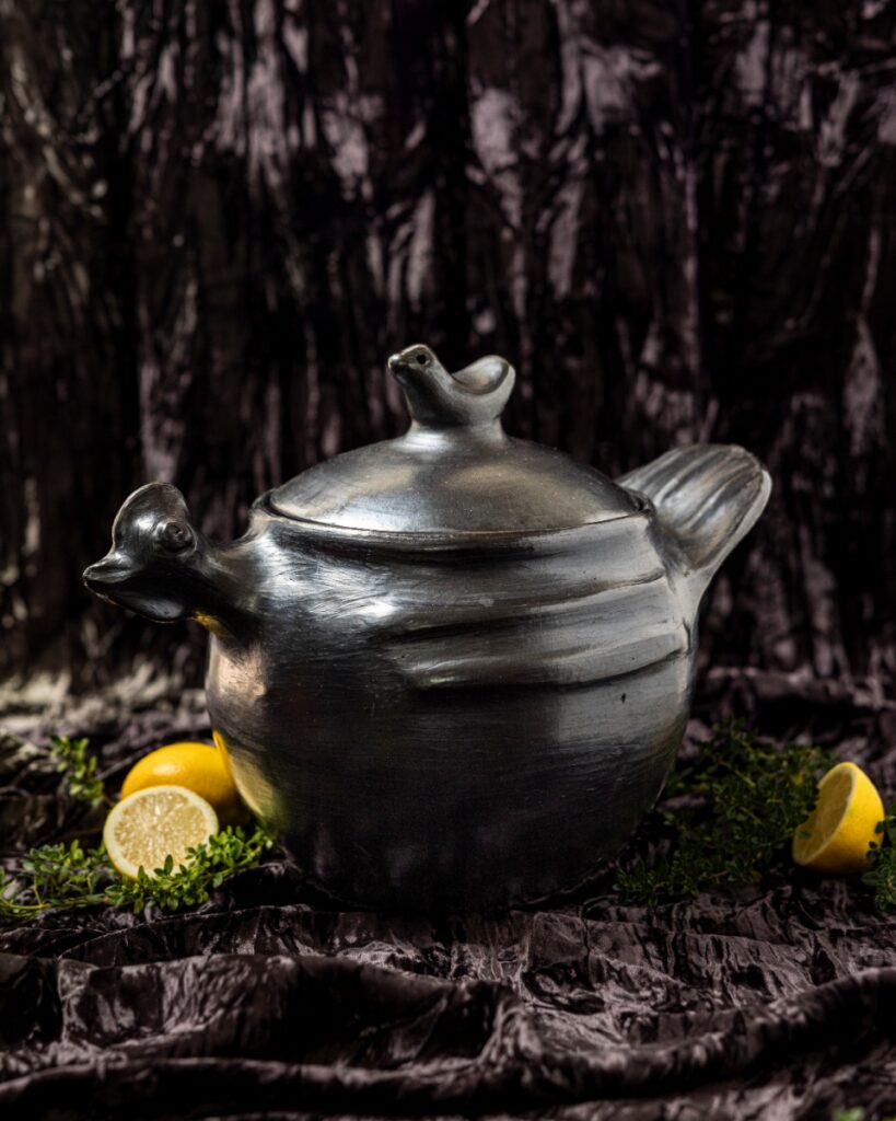 A black, burnished Chamba clay hen cooking pot with lid, surrounded by lemons and fresh thyme on a dark velvet surface.