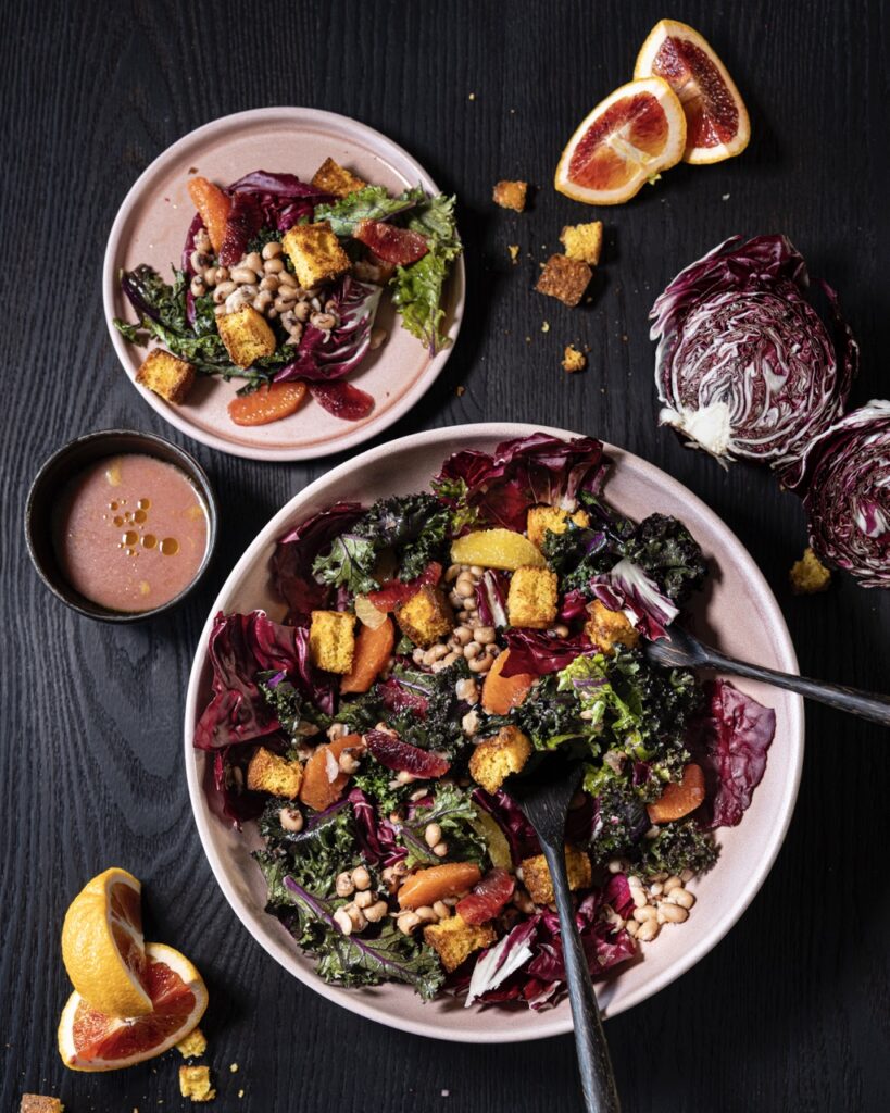 A big white bowl of salad with various toppings and black eyed peas beside a small pink plate with the salad on it.