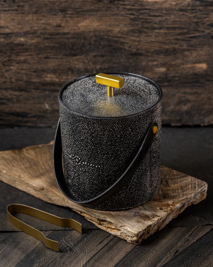 A black leather shagreen ice bucket with a brass accent handle on the lid, resting on a wooden plank with brass tongs.