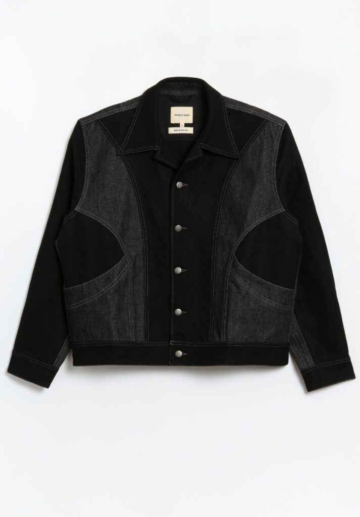 A black bomber jacket by Nigel Cabourn featuring gray denim contrast panels in a mid-century style with silver buttons.