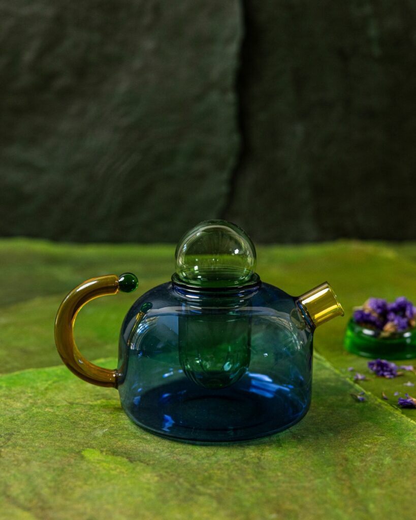 A small borosilicate glass teapot in a deep blue with an amber handle, on a bright green textured cloth.
