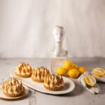 Four individual lemon meringue tarts with toasted meringue peaks, arranged on white platters next to fresh lemons and a white bust statue.