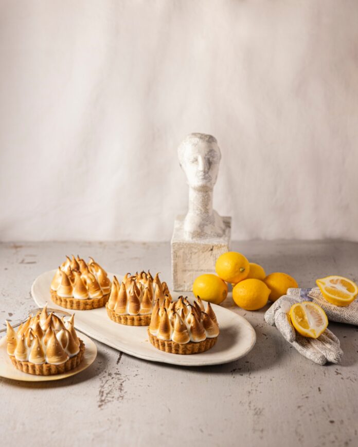 Four individual lemon meringue tarts with toasted meringue peaks, arranged on white platters next to fresh lemons and a white bust statue.