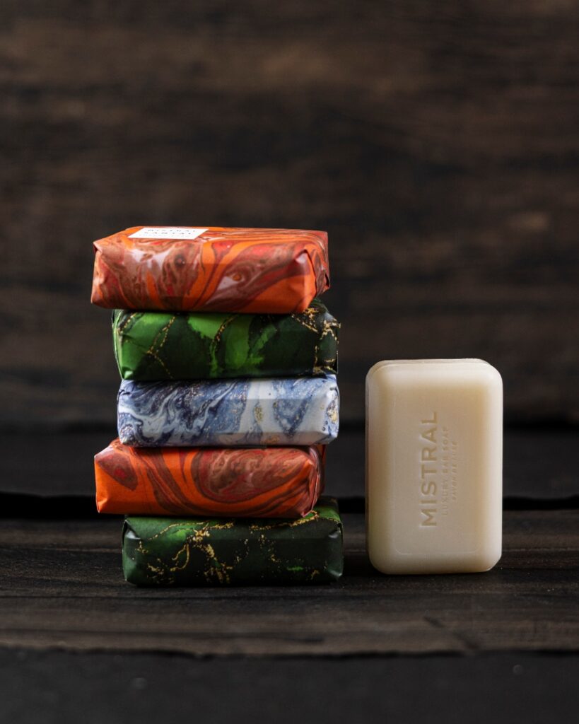 A stack of five brightly wrapped Mistral bar soaps next to an unwrapped pale bar, on a dark wooden table.