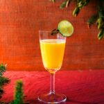A tall stemmed glass containing a vibrant orange Canelazo cocktail, garnished with a lime slice and cinnamon stick, set on a red wooden background with pine boughs.