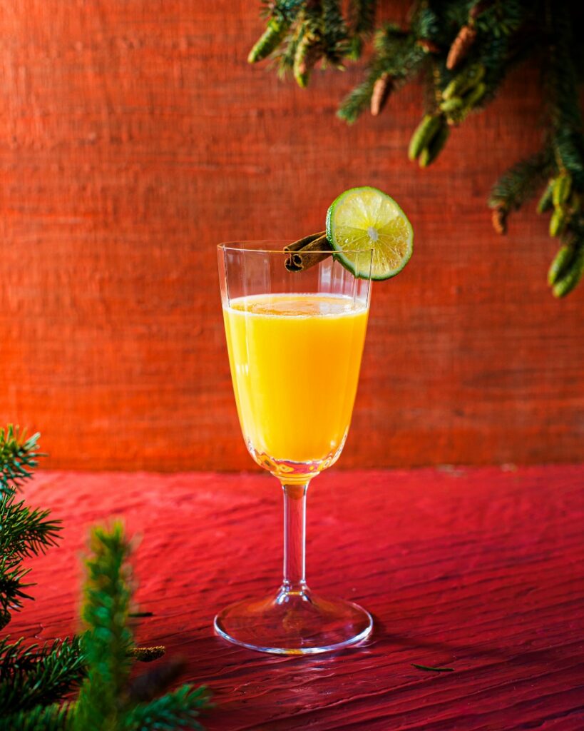 A tall stemmed glass containing a vibrant orange Canelazo cocktail, garnished with a lime slice and cinnamon stick, set on a red wooden background with pine boughs.