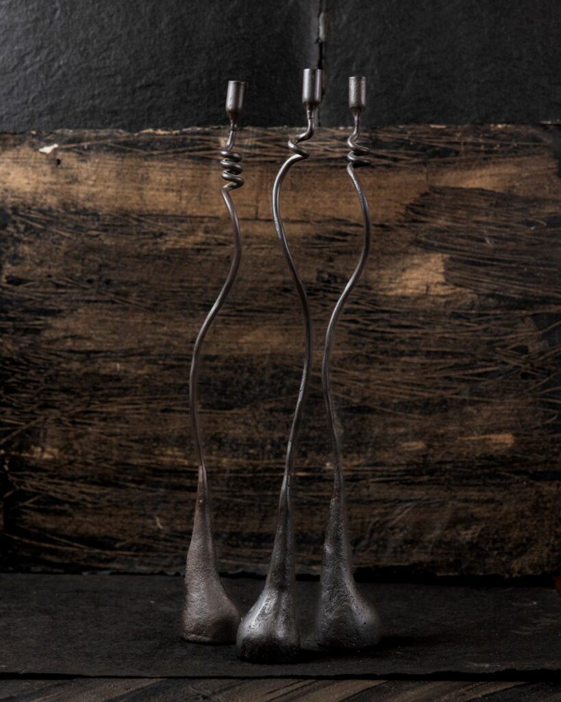 Three tall, slender, hand-forged iron Cats & Jasmine candle holders with swirling stems on a black textured background.