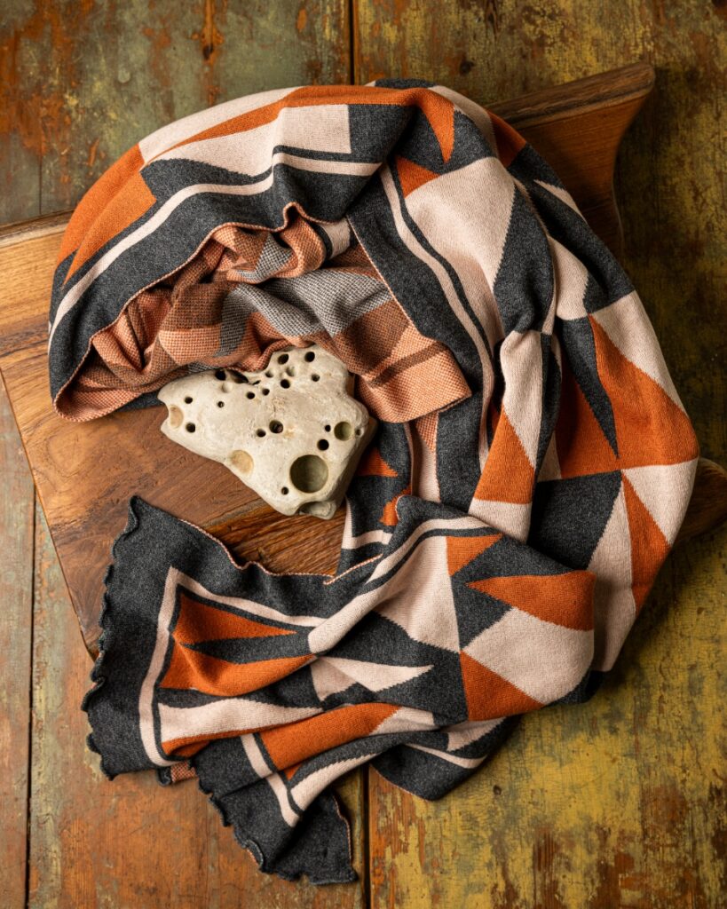 A charcoal and cinnamon geometric pattern cotton knit scarf from Liamolly draped over a wooden cutting board with a small, porous stone.