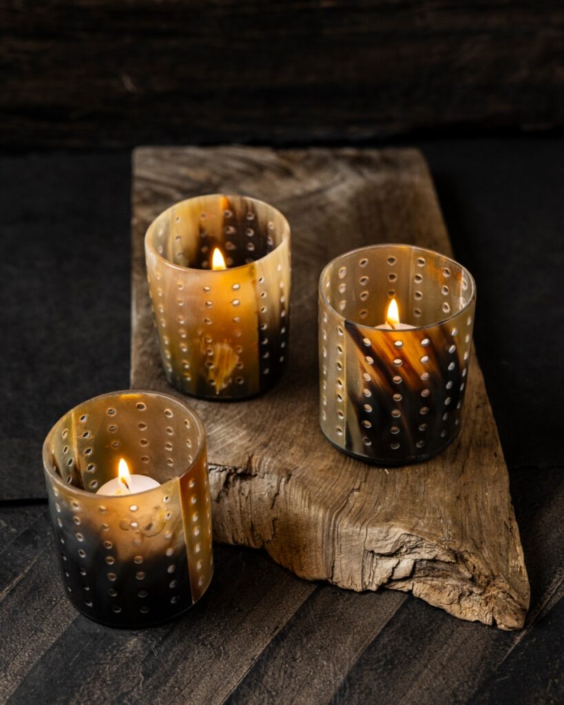 Three perforated tea light holders handcrafted in Kenya from natural, translucent Ankole-Watusi horn, glowing on a rustic wooden plank.