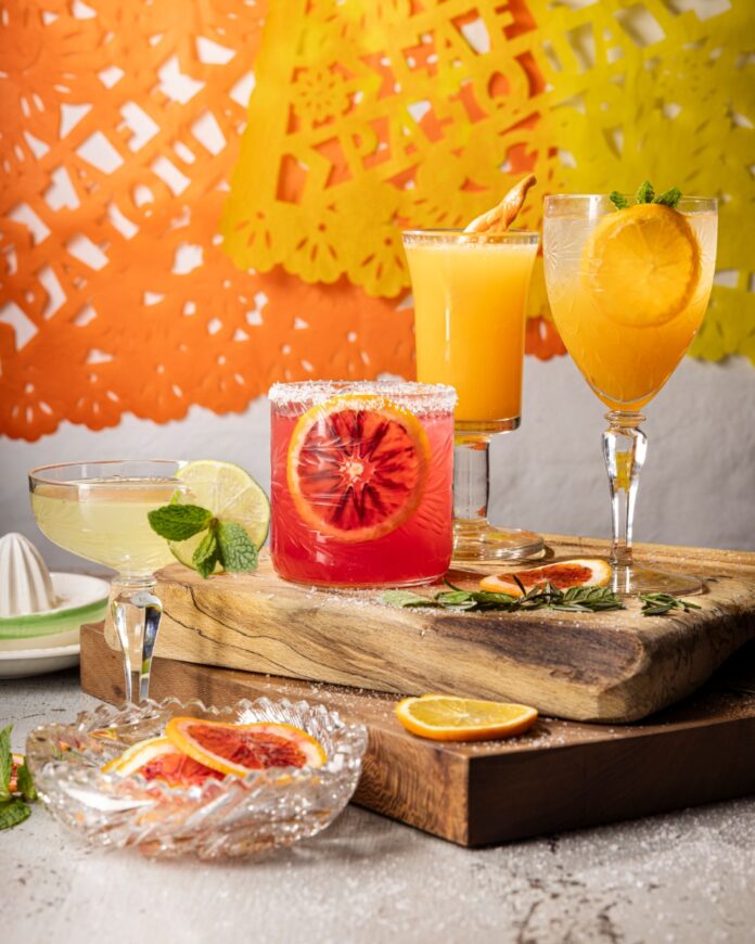 A dark red cocktail in a stemmed glass with a sugared rim and a slice of blood orange, flanked by two other citrus cocktails and festive cut paper decorations.