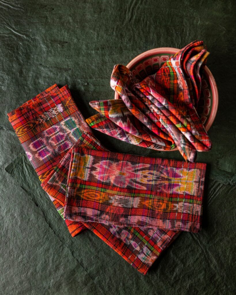 A stack of brightly colored, hand-woven ikat plaid napkins from Guatemala, with vibrant red, orange, and purple hues, resting on a dark green surface.
