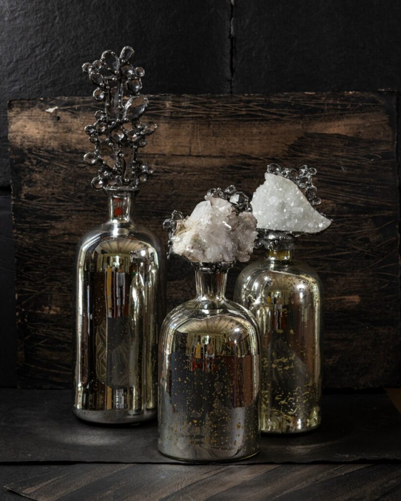 Three decorative mercury glass bottles with sculptural clear quartz crystal and metallic branch stoppers on a distressed wood and dark slate background.