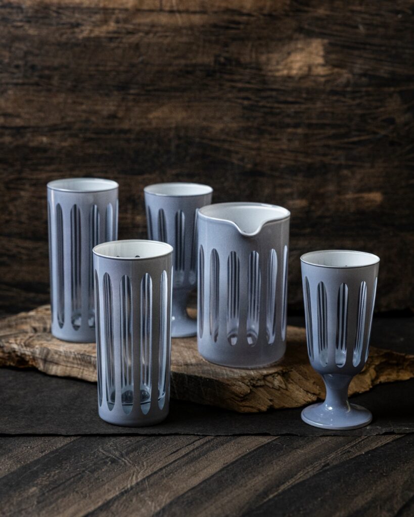 A set of five gray, footed Rialto drinking glasses and a pitcher with etched vertical lines, sitting on a weathered wooden surface.