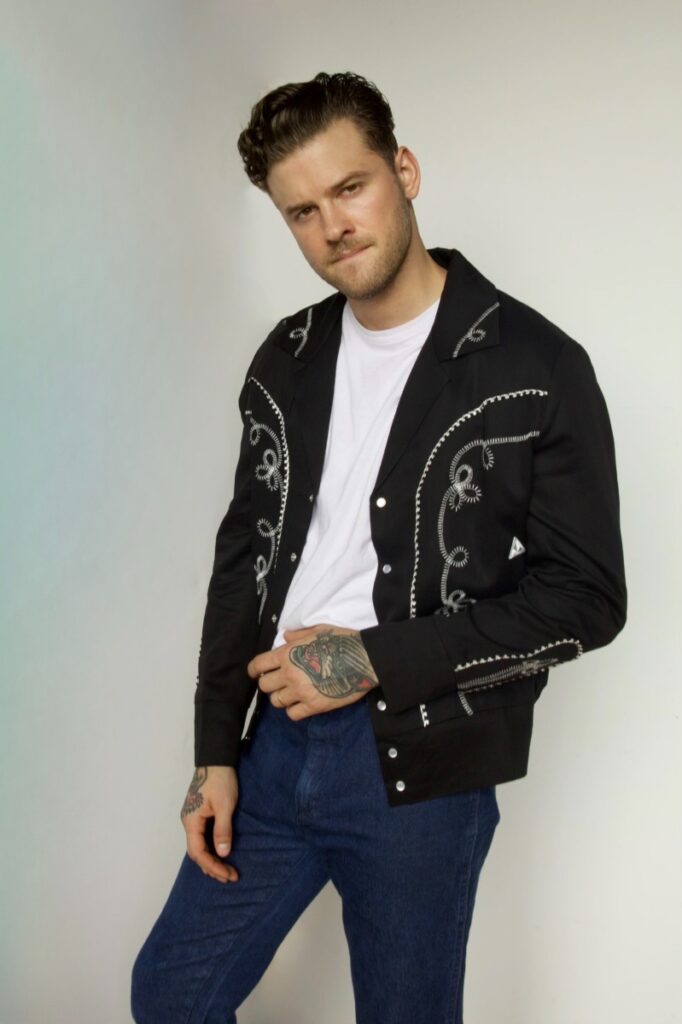 A man modeling a black H Bar C Western-style tencel jacket with white swirling embroidery, worn over a white t-shirt and blue jeans.